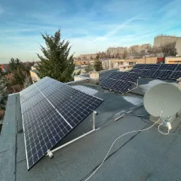 Instalacja fotowoltaiczna na płaskim dachu bloku mieszkalnego z anteną satelitarną w tle, widok na panoramę miasta pod błękitnym niebem.