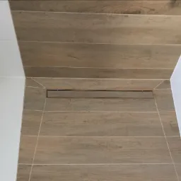 Ułożone poziomo płytki imitujące drewno w kabinie prysznicowej z odpływem liniowym, białe ściany po bokach.