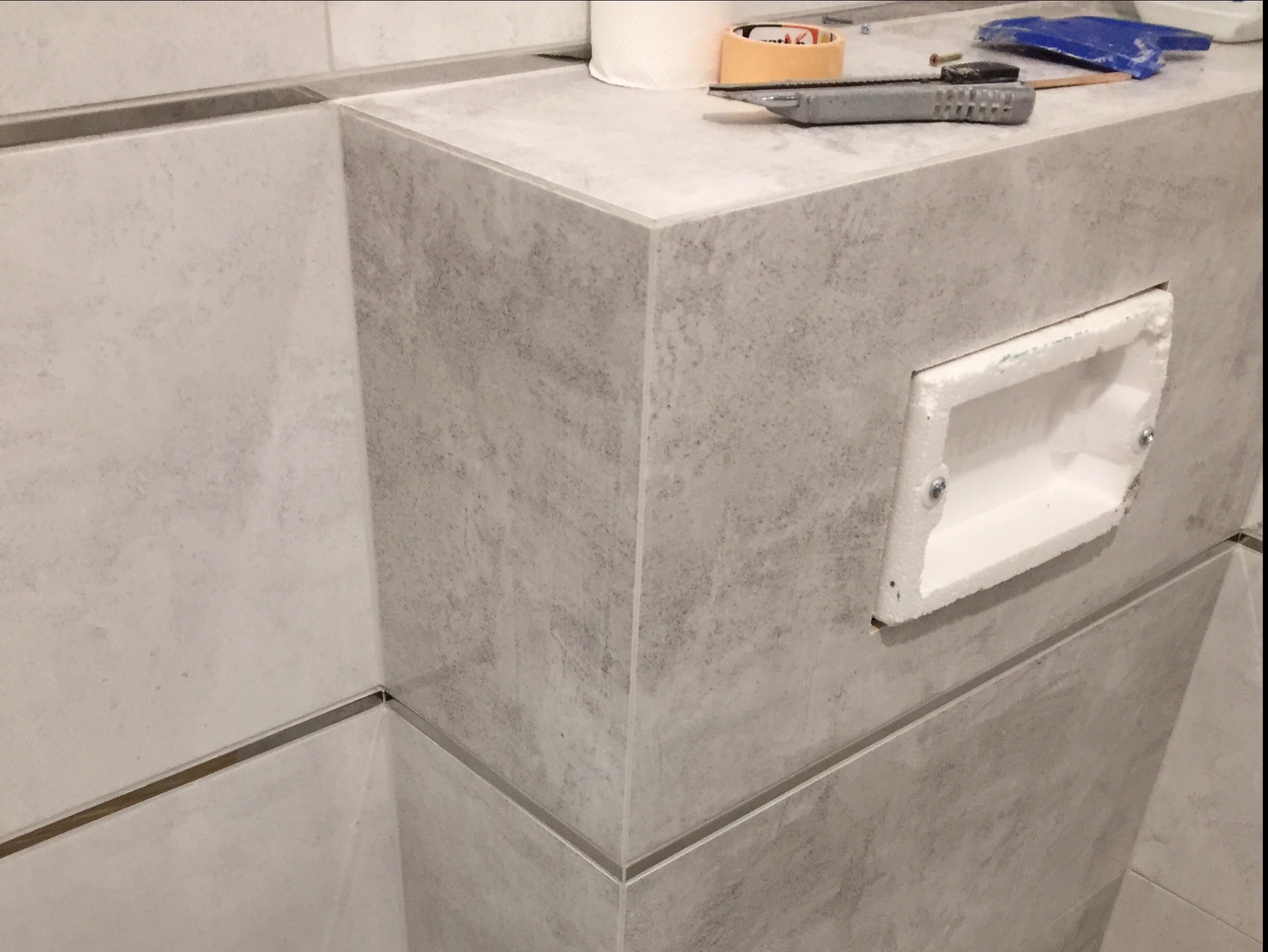 Szare płytki ceramiczne imitujące kamień, ułożone na ścianie i obudowie, z widocznym otworem na puszkę elektryczną, nożyk segmentowy i rolka taśmy malarskiej na górnej powierzchni.