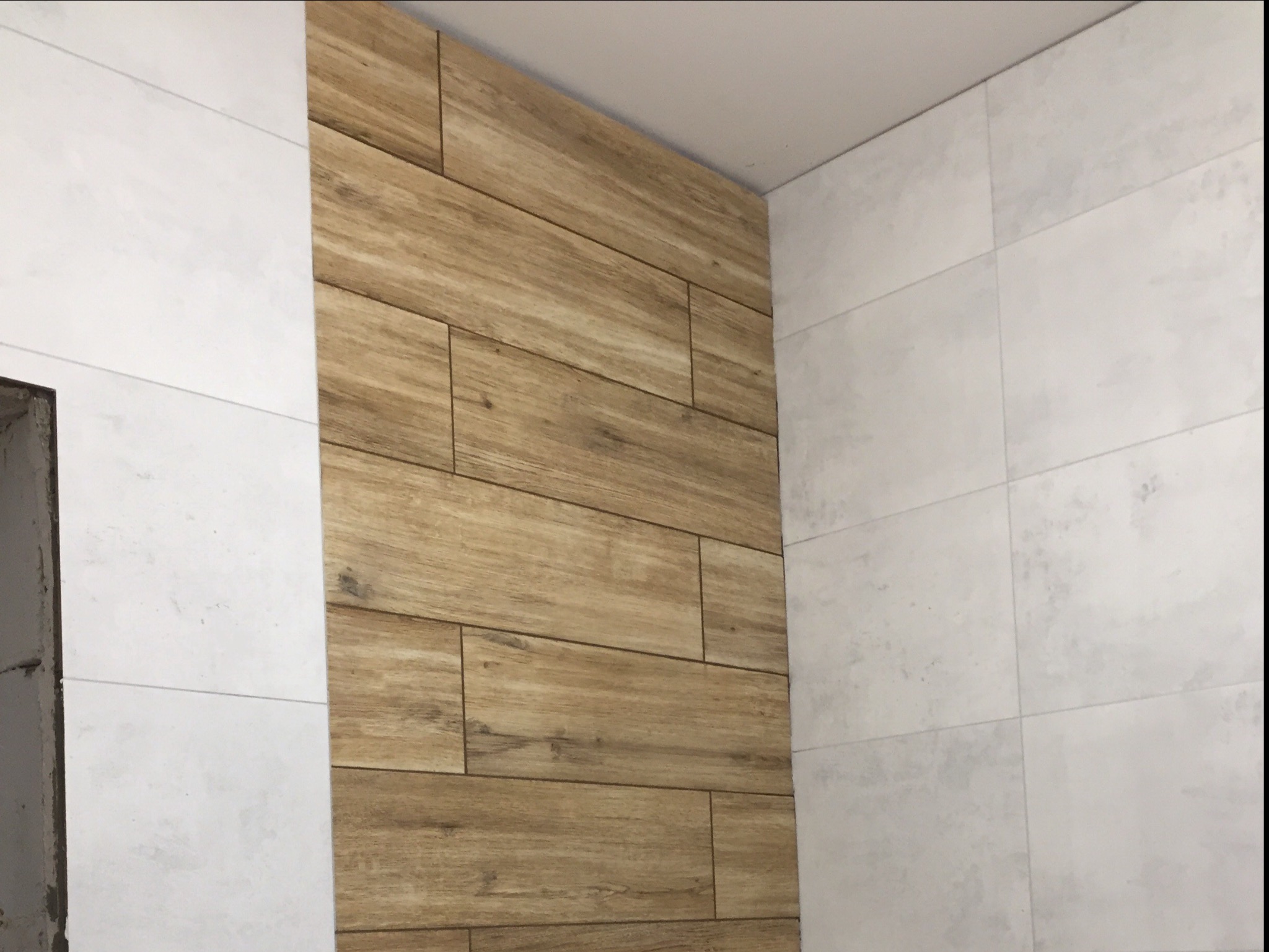 Ściana wykończona płytkami imitującymi drewno, sąsiadująca z białą ścianą wyłożoną płytkami o strukturze betonu w łazience.