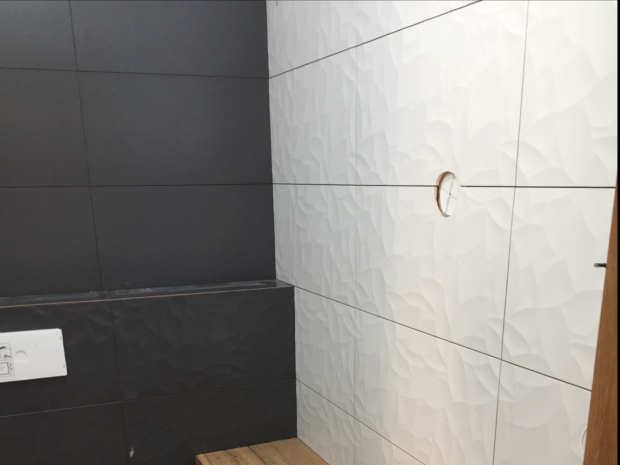 Częściowo wykończona ściana w łazience z połączeniem ciemnoszarych i białych płytek z fakturą fal, otwór na instalację sanitarną w białych płytkach.