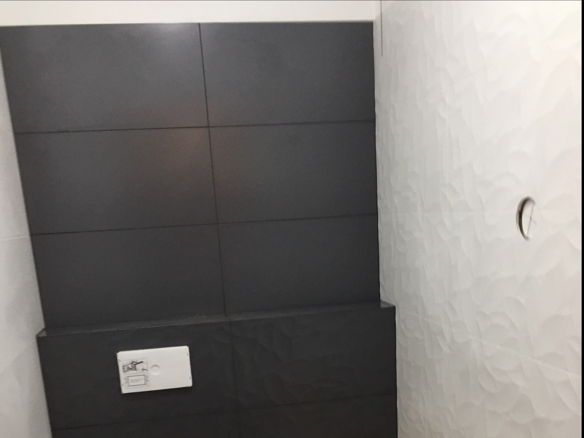 Częściowo wykończona łazienka z szarymi płytkami na ścianie obudowy toalety i białymi płytkami z falistą teksturą na pozostałej części ściany, widoczny przycisk spłuczki.