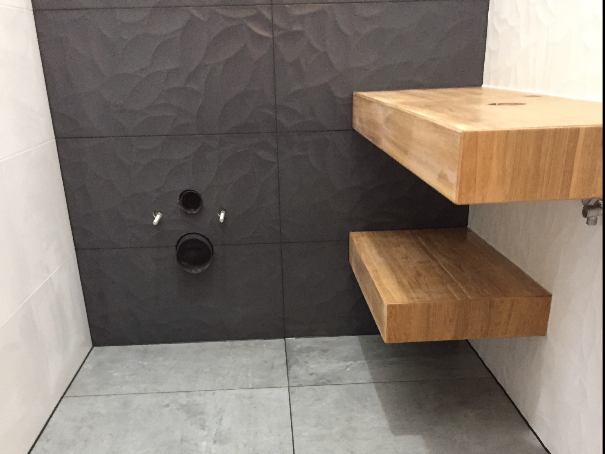 Łazienka w trakcie wykańczania: ciemnoszare, strukturalne płytki na ścianie z widocznymi otworami instalacyjnymi, jasne, drewniane półki zamontowane na ścianie, szare płytki podłogowe.