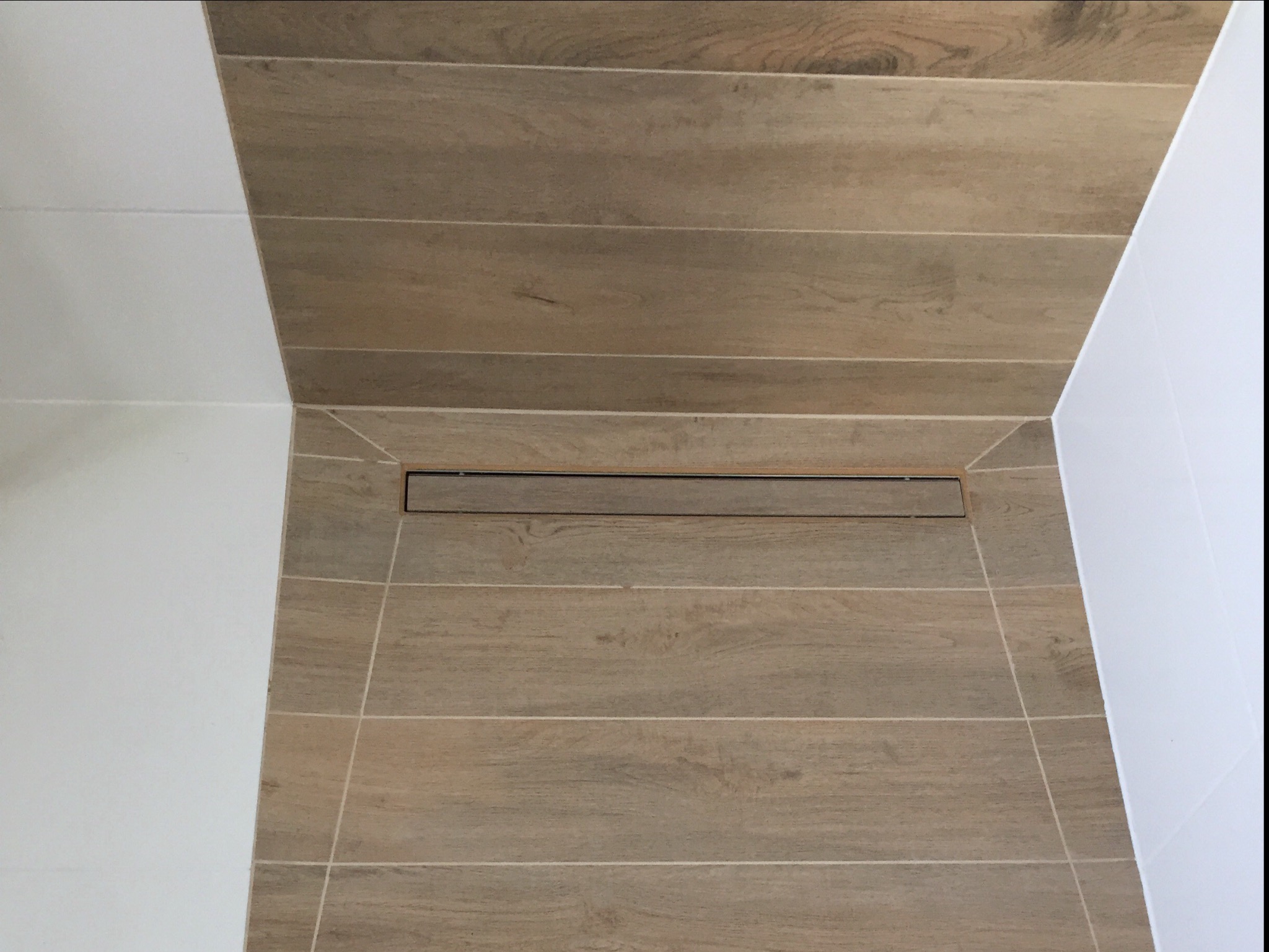 Ułożone poziomo płytki imitujące drewno w kabinie prysznicowej z odpływem liniowym, białe ściany po bokach.