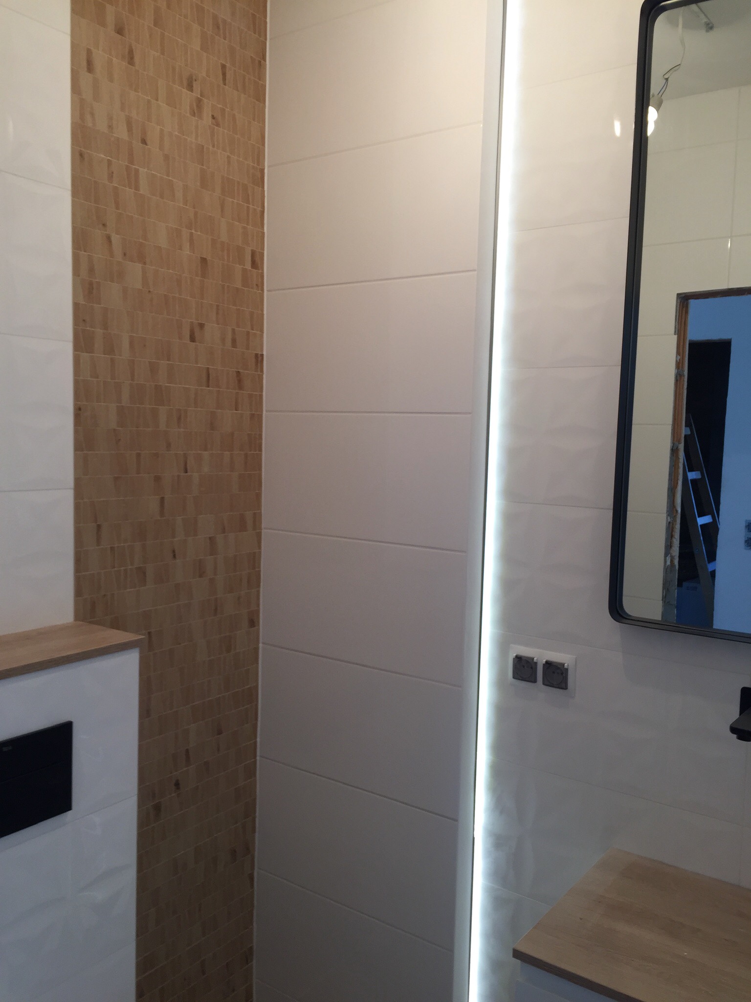 Wykończona łazienka z białymi płytkami ściennymi, pionowym pasem płytek imitujących drewno, podświetlanym lustrem w czarnej ramie i drewnianym blatem umywalki.