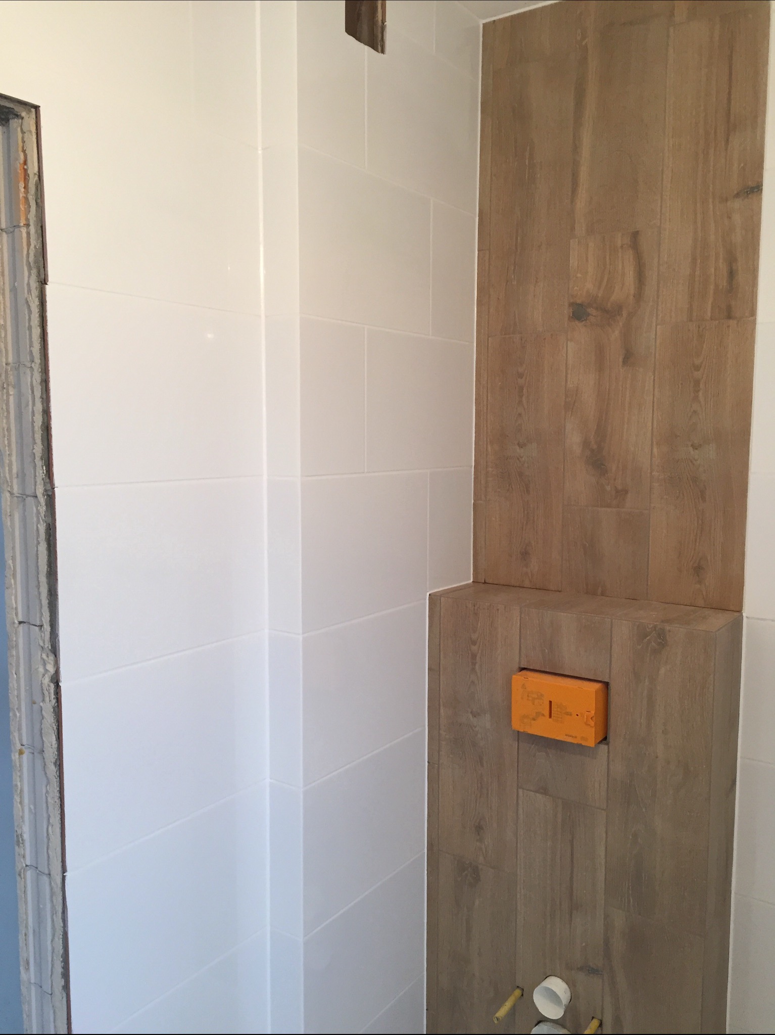 Fragment łazienki w trakcie remontu: białe płytki na ścianie sąsiadują z drewnopodobną okładziną, widoczna puszka instalacyjna i fragment surowego ościeża drzwi.