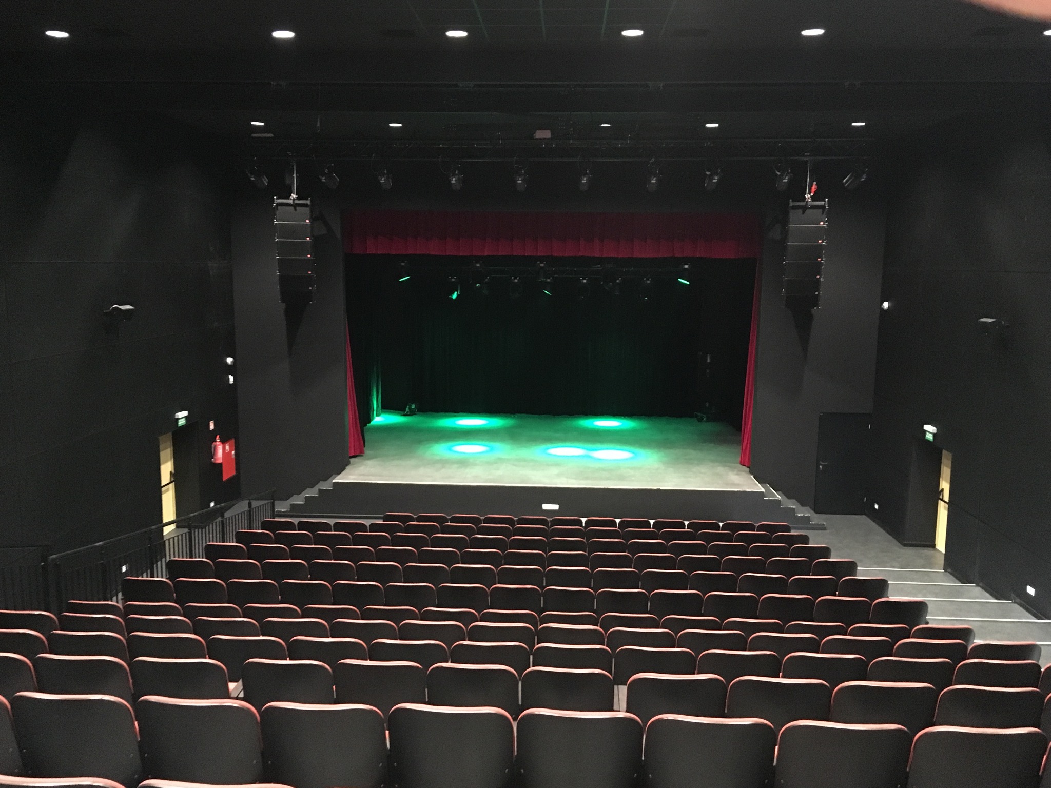 Widok z góry na salę widowiskową z rzędami foteli, sceną z czerwonymi kurtynami i oświetleniem punktowym w kolorze zielonym, z widocznymi głośnikami po bokach sceny.