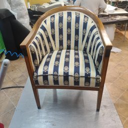 F. H Franco - Odnowiony fotel z drewnianą ramą i tapicerką w niebiesko-złote pasy, stojący na metalowym stole w warsztacie. W tle widoczne materiały i osoby pracujące.