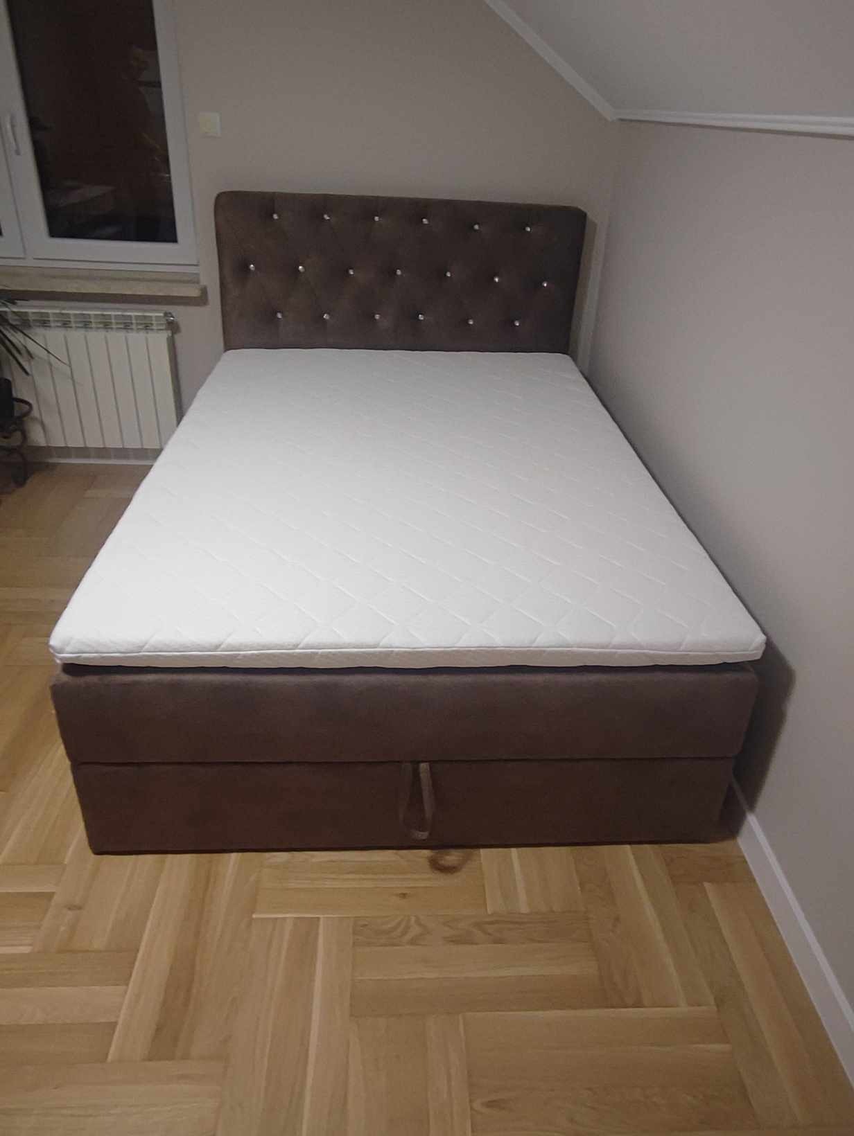 Tapicerowane łóżko z brązowym wezgłowiem zdobionym kryształkami, materacem i pojemnikiem na pościel, ustawione na parkiecie w jasnym pokoju.