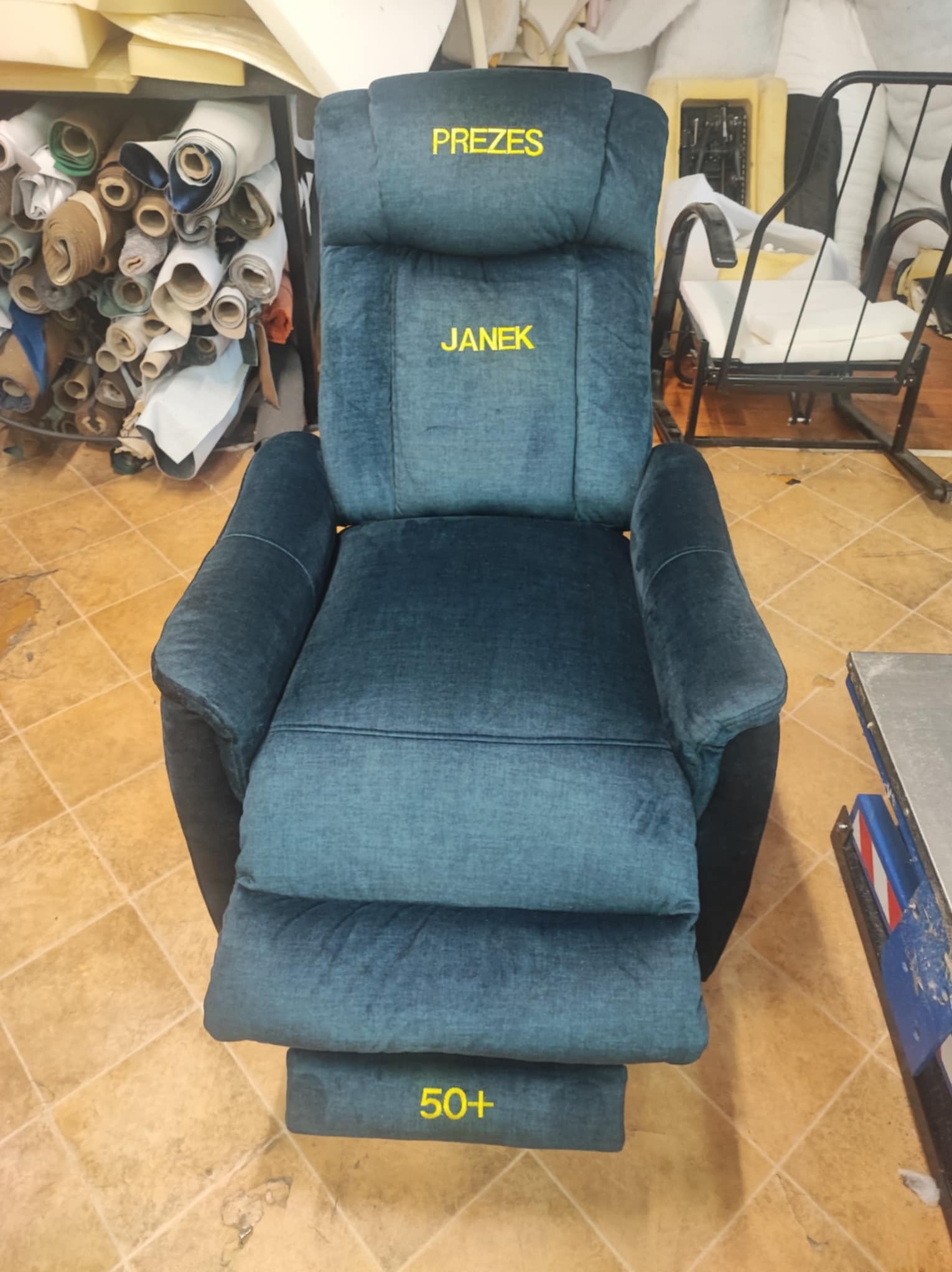Fotel z nową, niebieską tapicerką, z napisami 'PREZES', 'JANEK' i '50+', stojący w warsztacie tapicerskim z widocznymi materiałami i narzędziami.