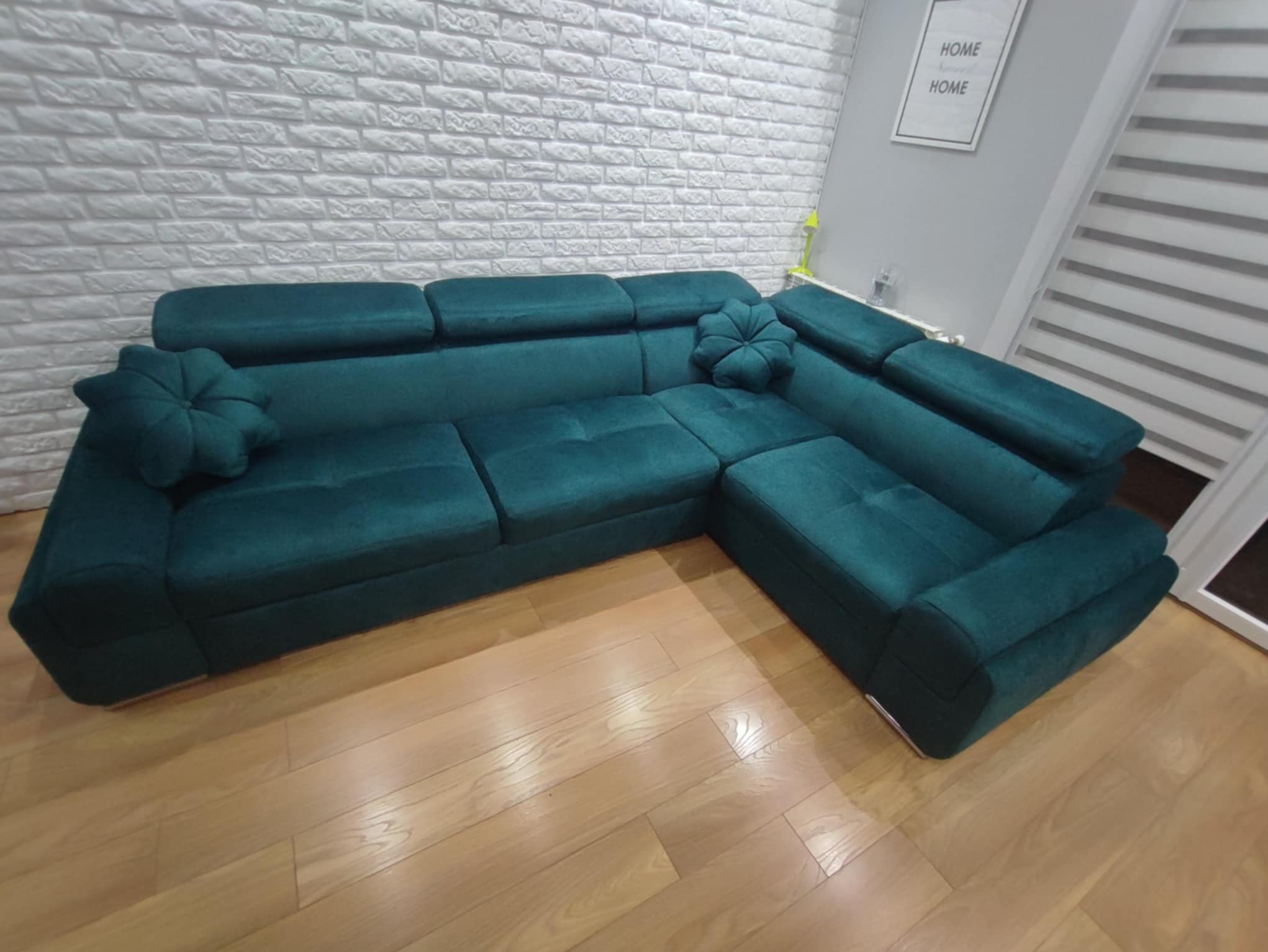 Szmaragdowa sofa narożna z poduszkami na drewnianej podłodze, na tle białej ściany z cegły. Widoczne detale tapicerki i dekoracyjne poduszki w kształcie kwiatów.