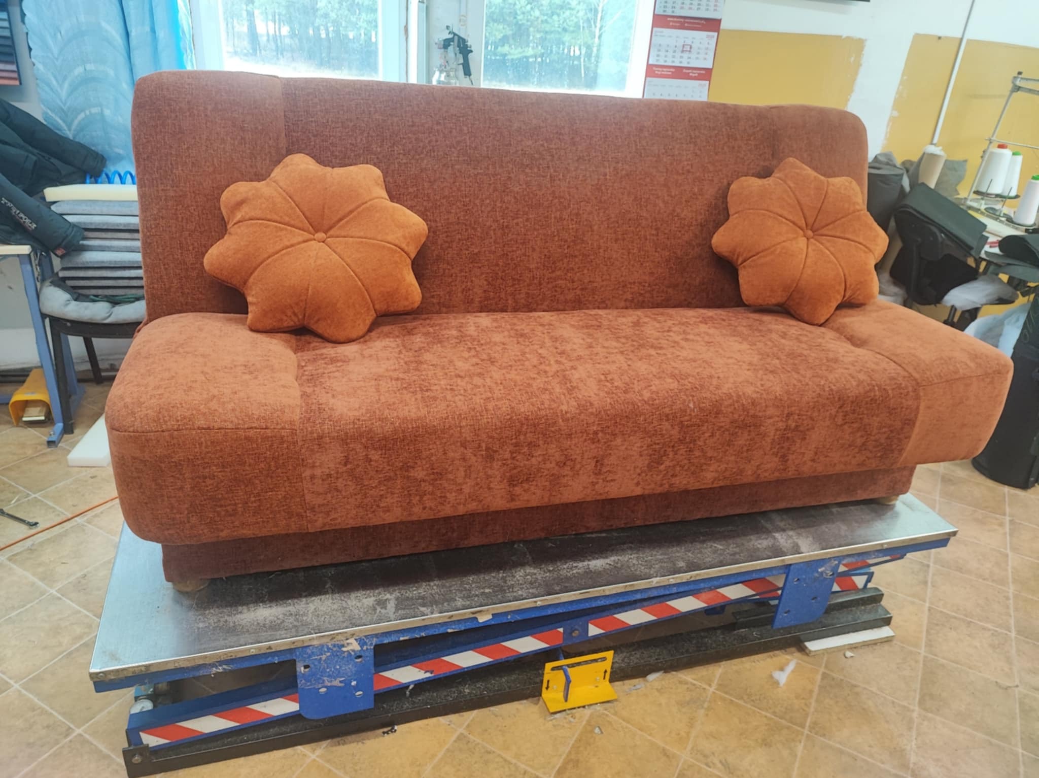 Brązowa sofa z dekoracyjnymi poduszkami w kształcie kwiatów, umieszczona na podnośniku w warsztacie. Widoczne materiały i narzędzia tapicerskie w tle.