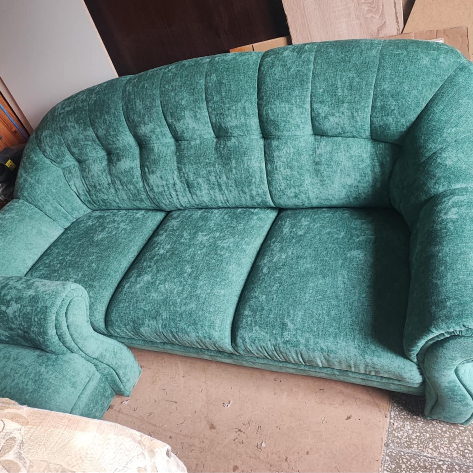 Turkusowa sofa z grubym obiciem w stylu retro. Widoczne marszczenia i zaokrąglone podłokietniki. Sofa stoi na brązowej podłodze w pomieszczeniu.