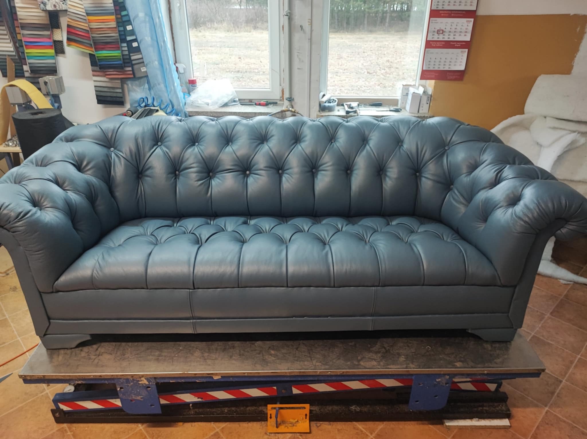 Niebieska sofa Chesterfield z pikowaniem na tle okna i wzorników tkanin, na podnośniku warsztatowym. Widoczne narzędzia i kalendarz.