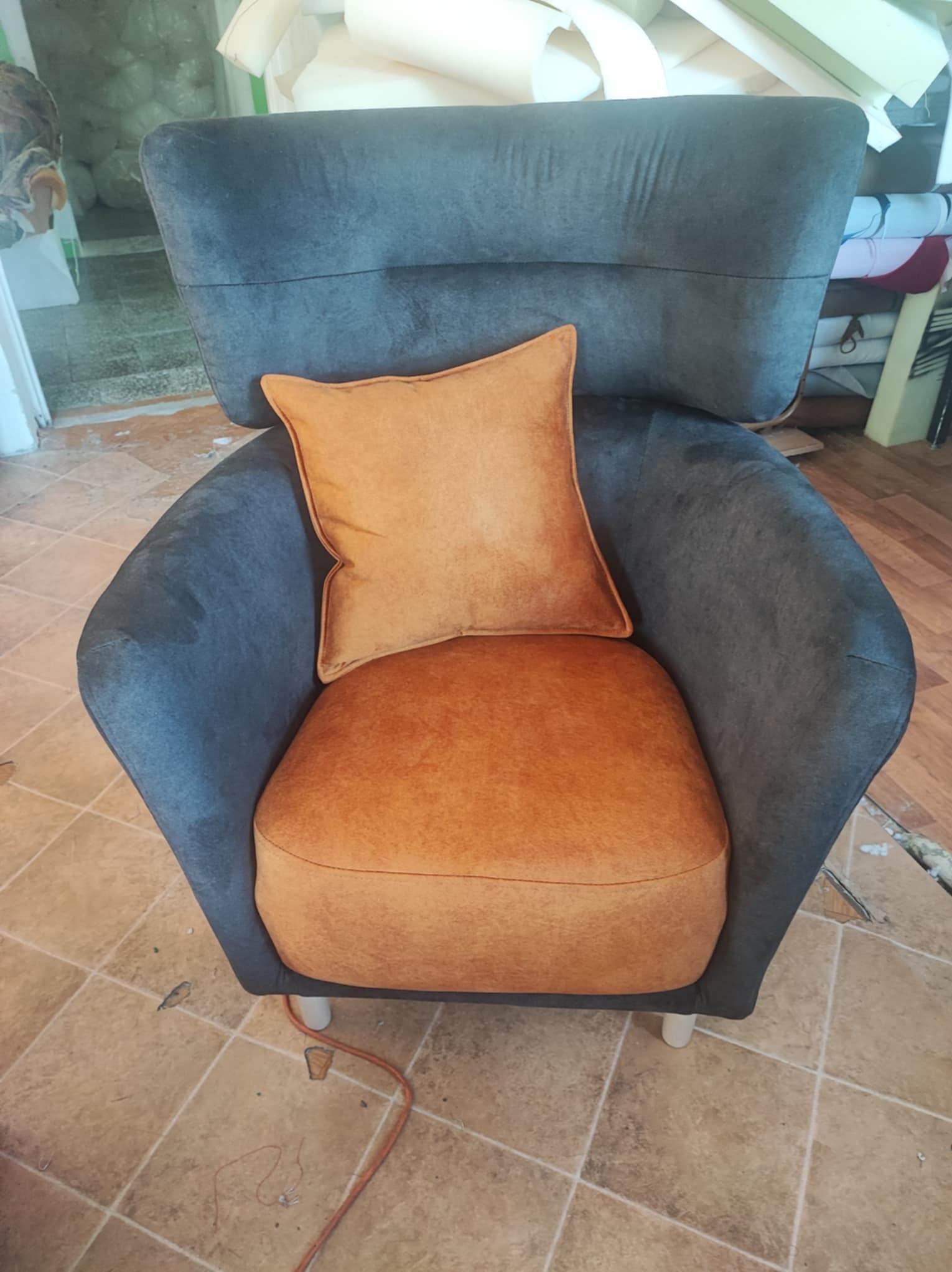 Odnowiony fotel w odcieniach szarości i pomarańczy z poduszką, stojący na kafelkach. Widoczne ślady pracy w tle, sugerujące warsztat.