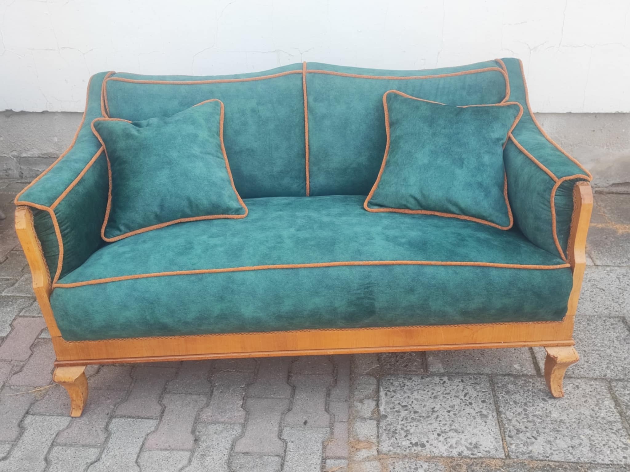 Dwuosobowa sofa w stylu retro, z zieloną tapicerką i pomarańczowymi lamówkami, na tle białej ściany i szarej kostki brukowej. Widoczne drewniane nóżki.