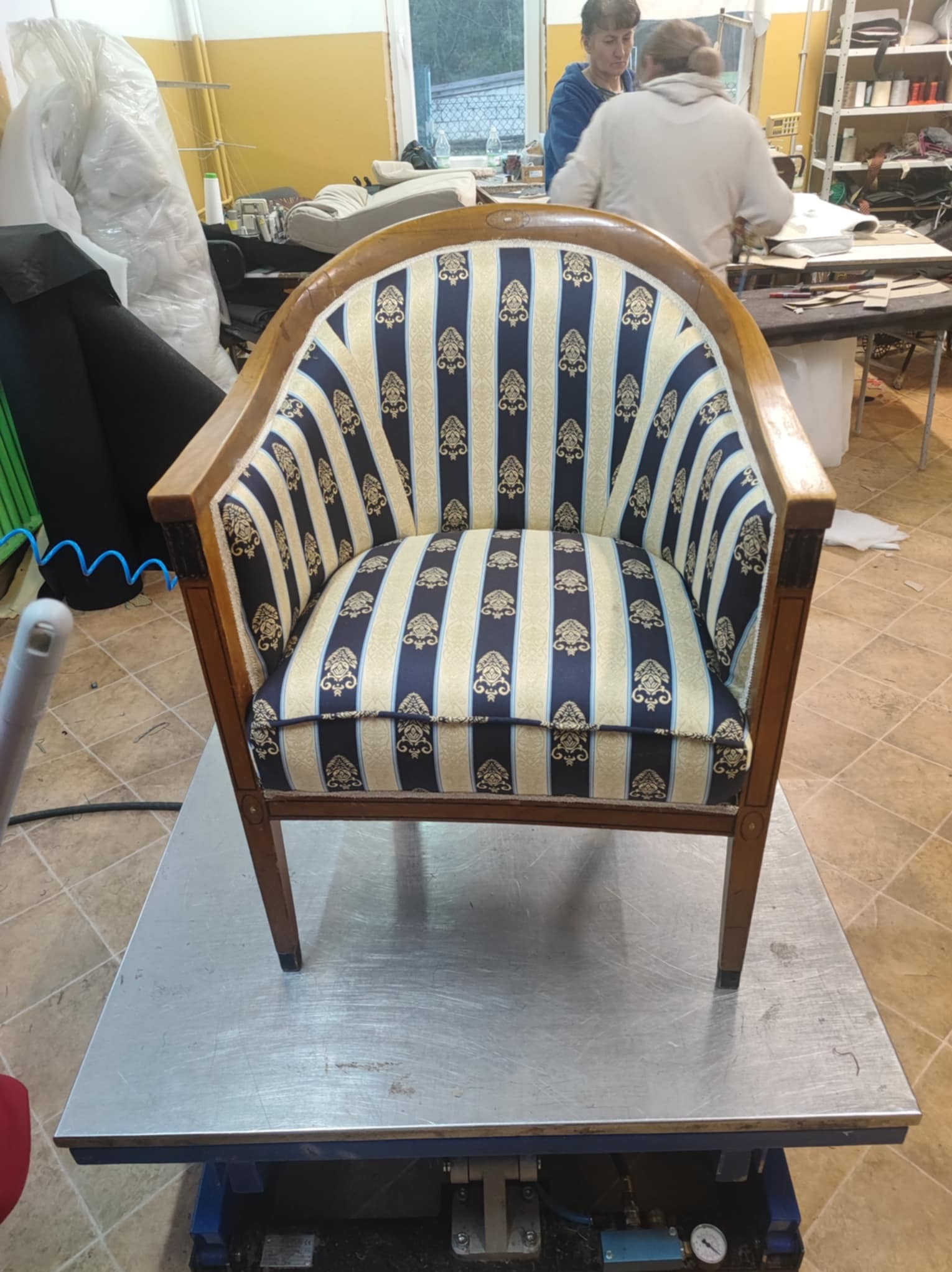 Odnowiony fotel z drewnianą ramą i tapicerką w niebiesko-złote pasy, stojący na metalowym stole w warsztacie. W tle widoczne materiały i osoby pracujące.