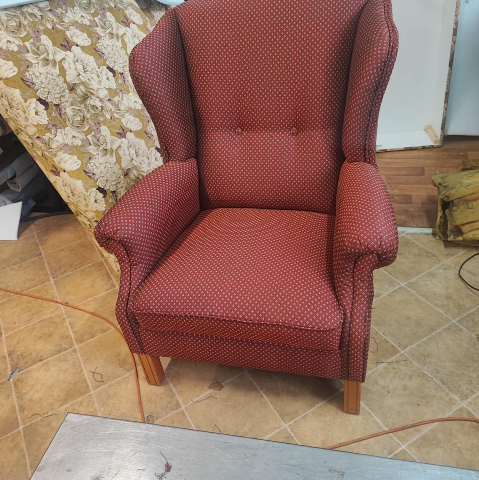 Czerwony fotel uszak z subtelnym wzorem w pracowni, obok widoczna stara tapicerka z motywem kwiatowym, sugerująca proces renowacji mebla.