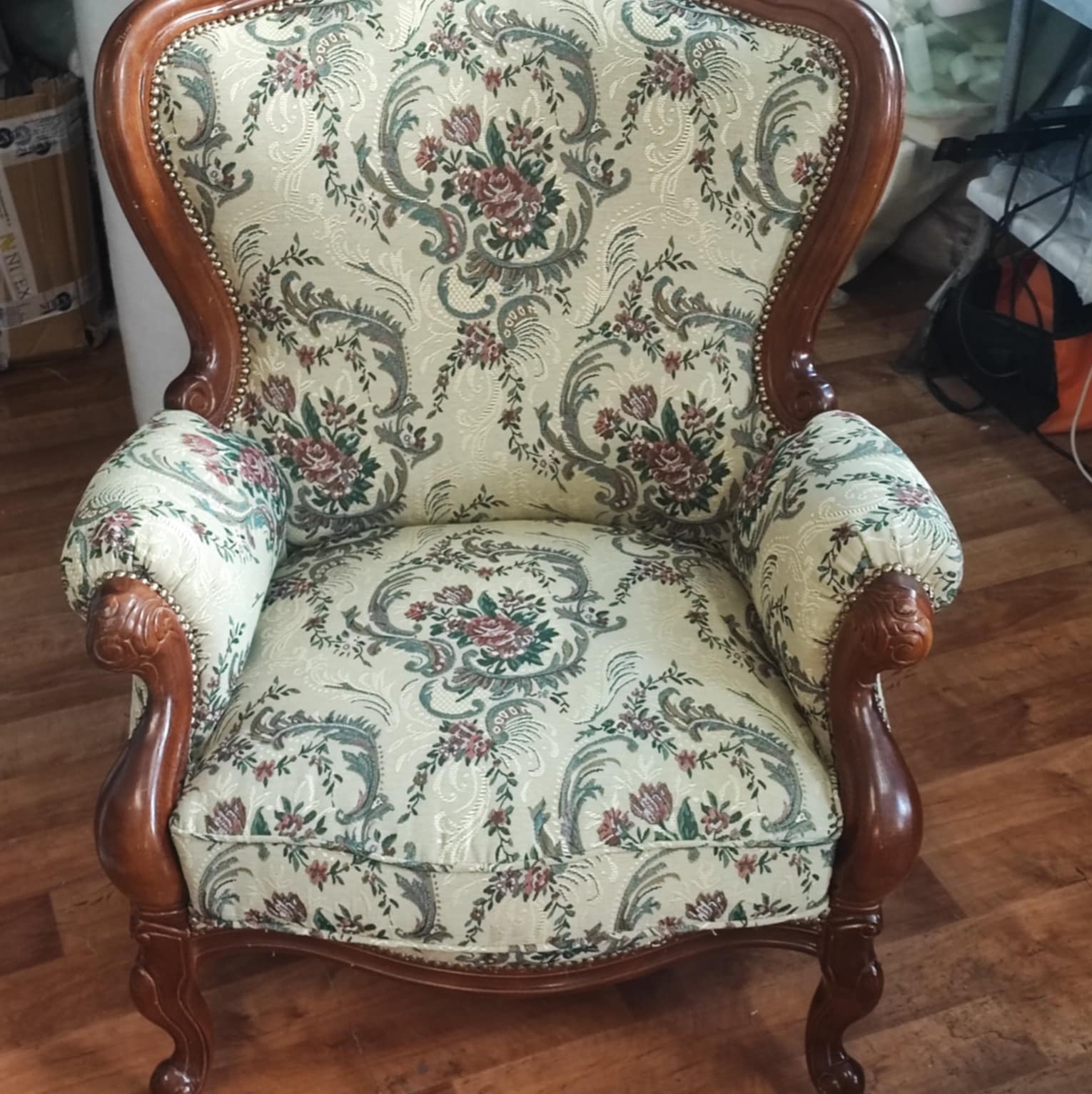 Stylowy fotel z drewnianą ramą i kremową tapicerką w kwiatowe wzory, widoczny na tle drewnianej podłogi, z detalami wykończenia podkreślającymi elegancję.