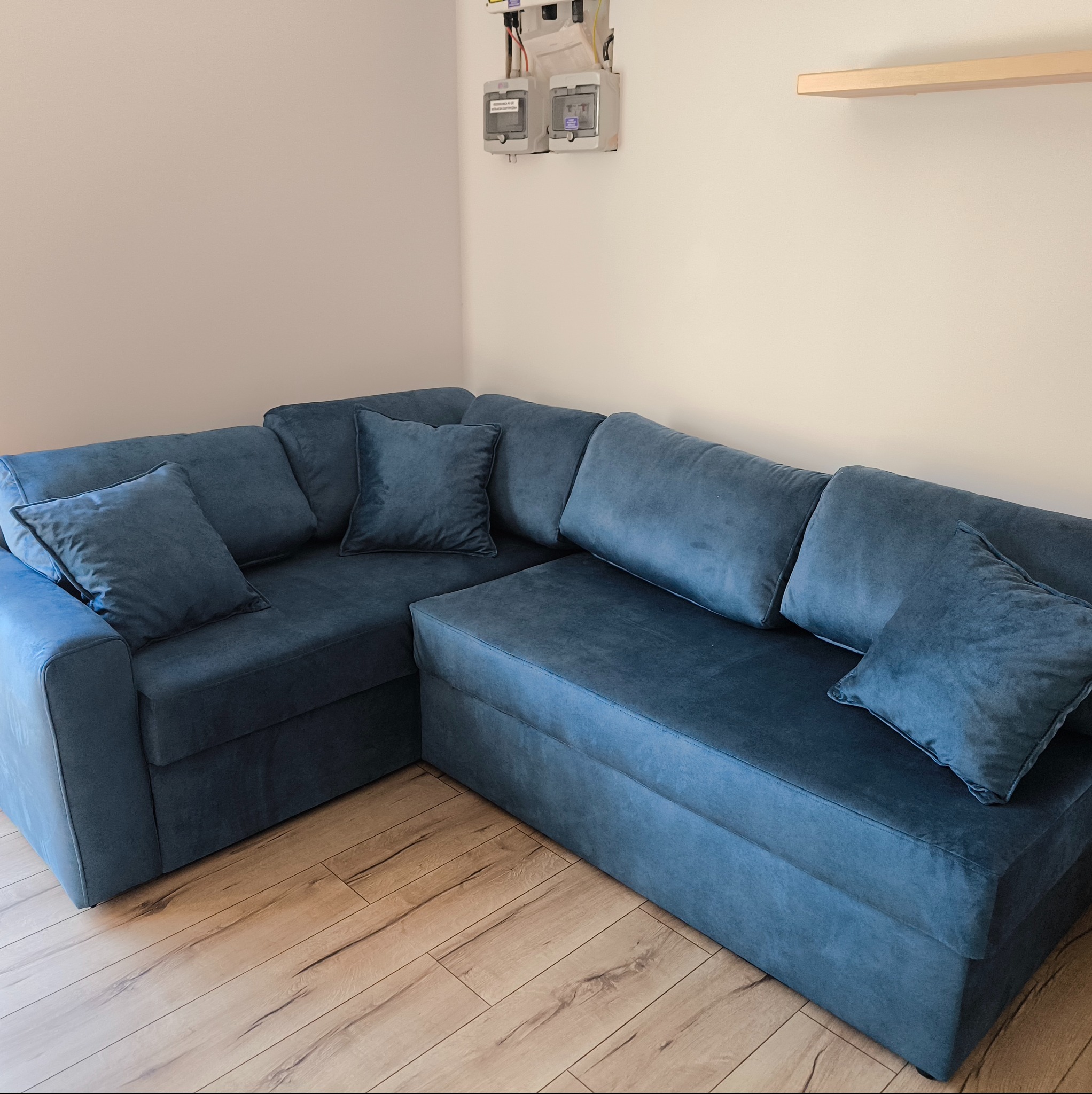 Narożnik w odcieniach niebieskiego z poduszkami, na drewnianej podłodze, w jasnym wnętrzu. Nad nim widoczne liczniki elektryczne i półka.