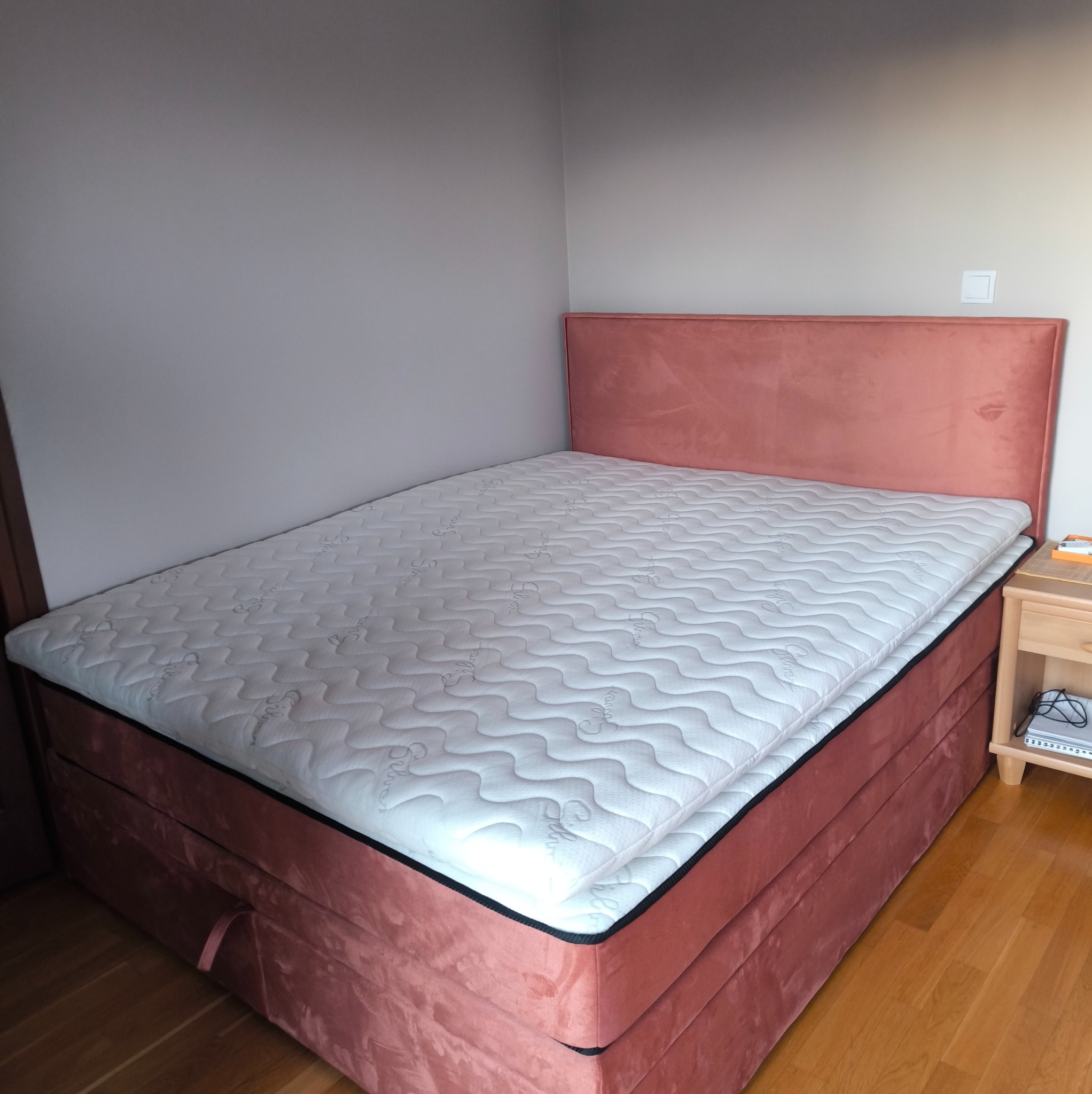 Różowe, tapicerowane łóżko kontynentalne z materacem, widoczne na tle szarej ściany i drewnianej podłogi. Obok szafka nocna z książkami.