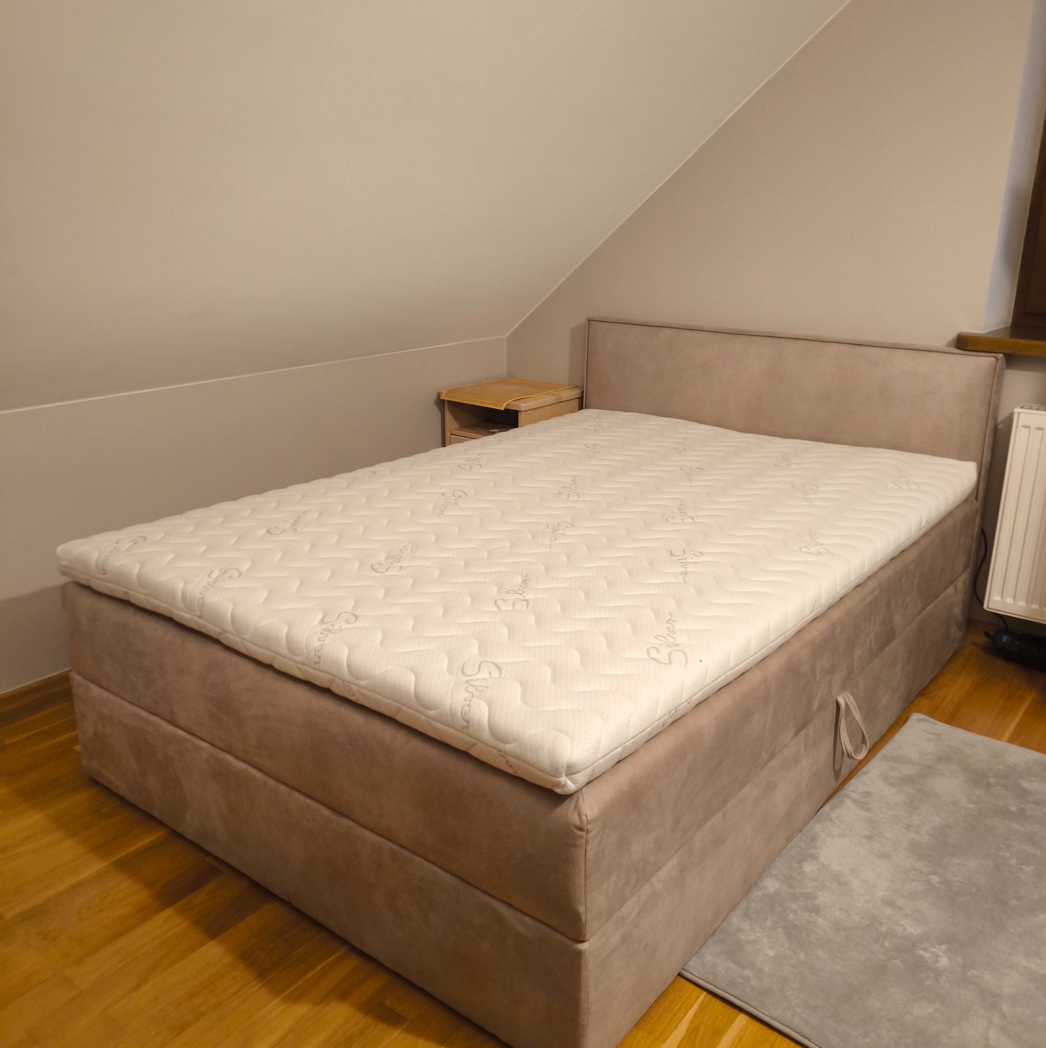 Tapicerowane łóżko kontynentalne w beżowym odcieniu, z materacem nawierzchniowym w jasnym pokoju na poddaszu. Widoczny fragment dywanu i szafki nocnej.
