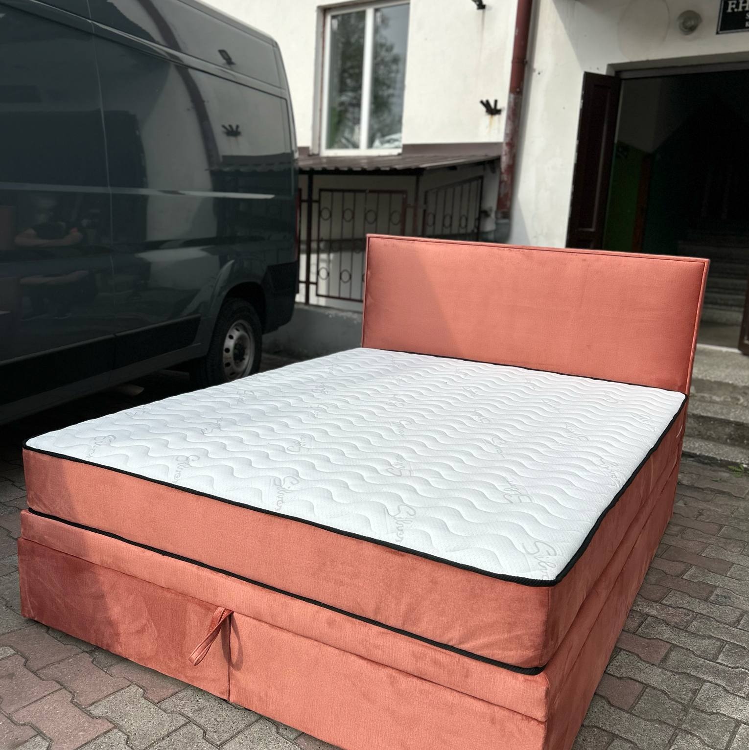 Łóżko tapicerowane w odcieniach koralowych z białym materacem na zewnątrz, obok szarego vana. Widoczny detal wykończenia i faktura materiału.