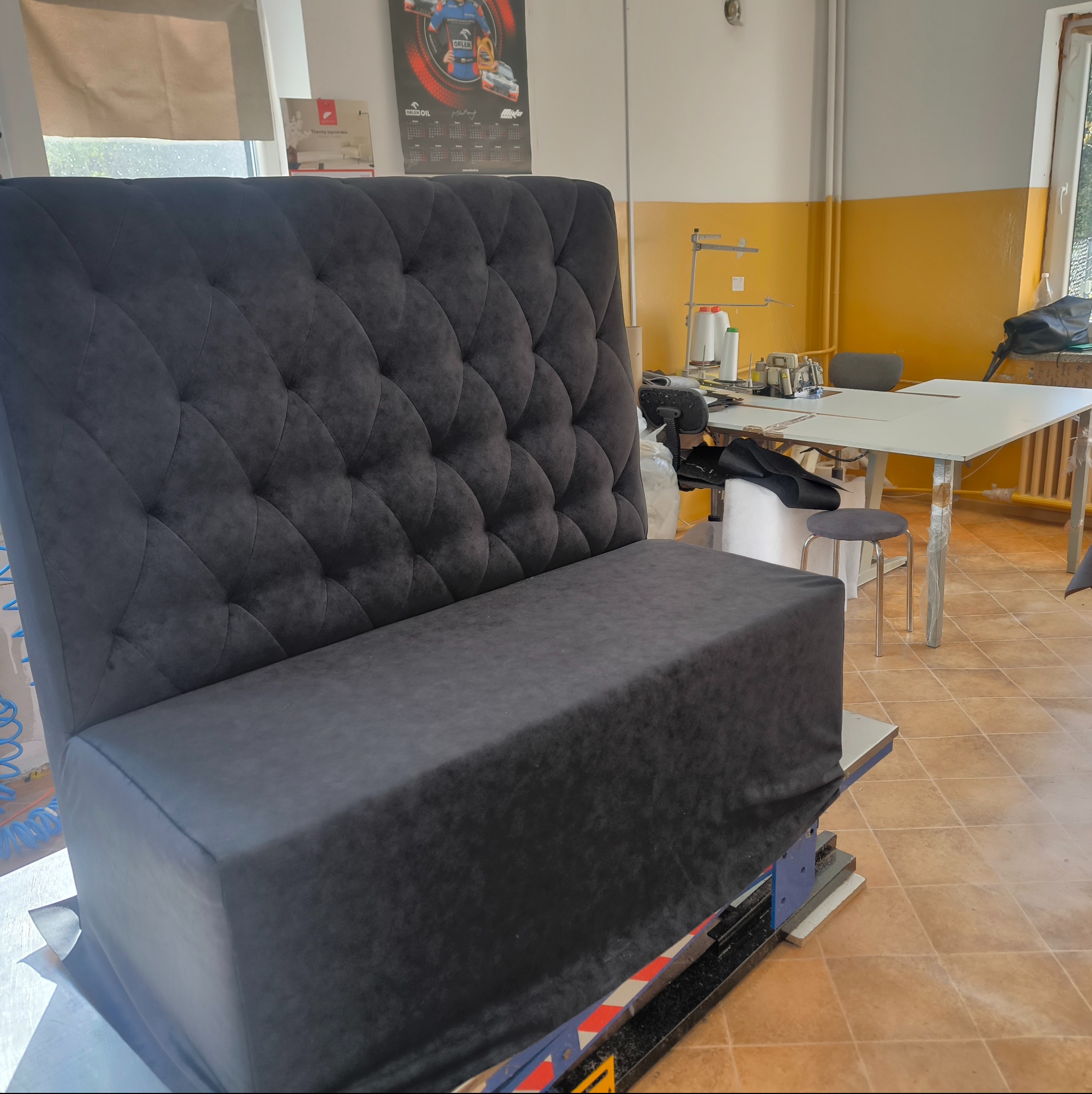 Ciemnoszara, pikowana ławka w warsztacie, widoczna maszyna do szycia w tle. Gotowy mebel z charakterystycznym wzorem pikowania.