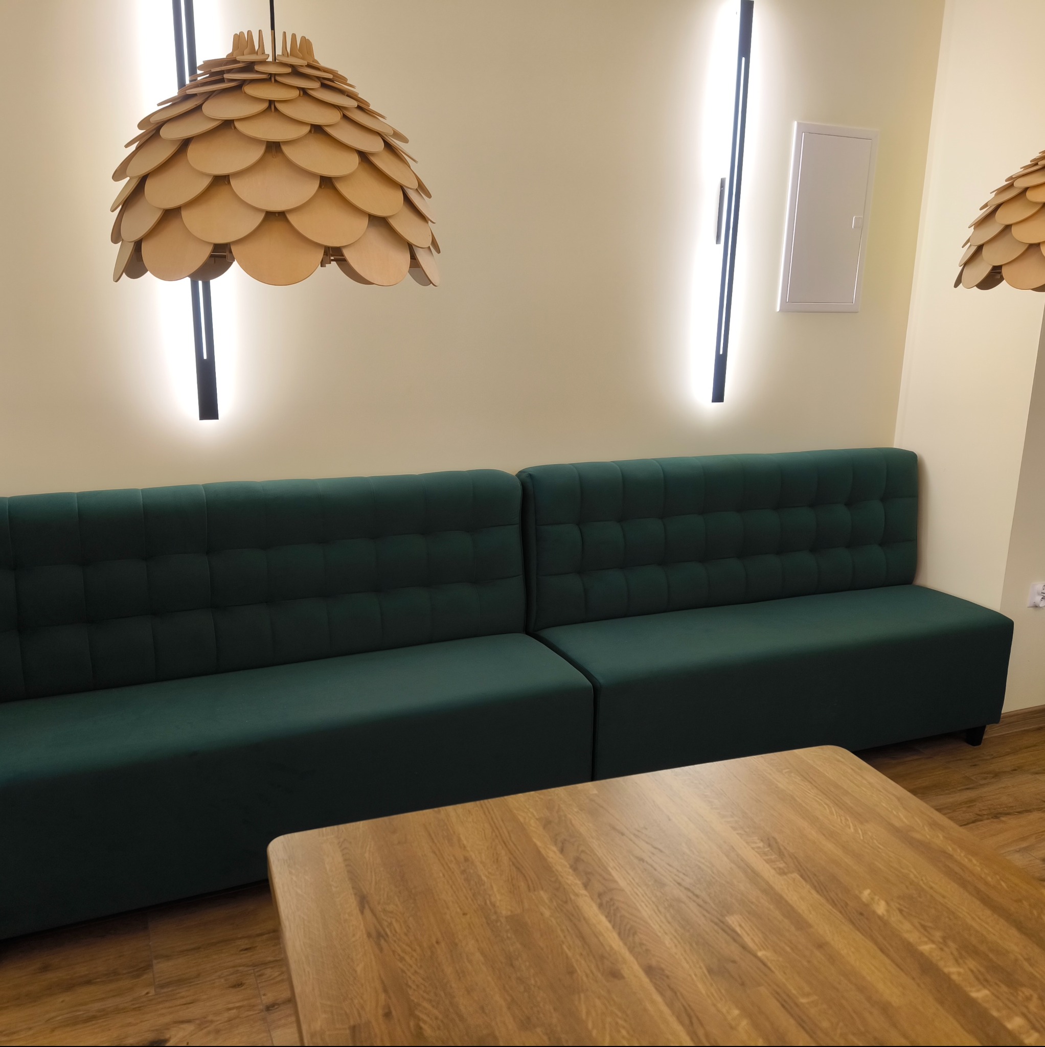 Zielona, pikowana sofa w lokalu we Frampolu. Drewniany stół i lampy z drewnianych krążków. Nowoczesne oświetlenie LED. Wnętrze lokalu.