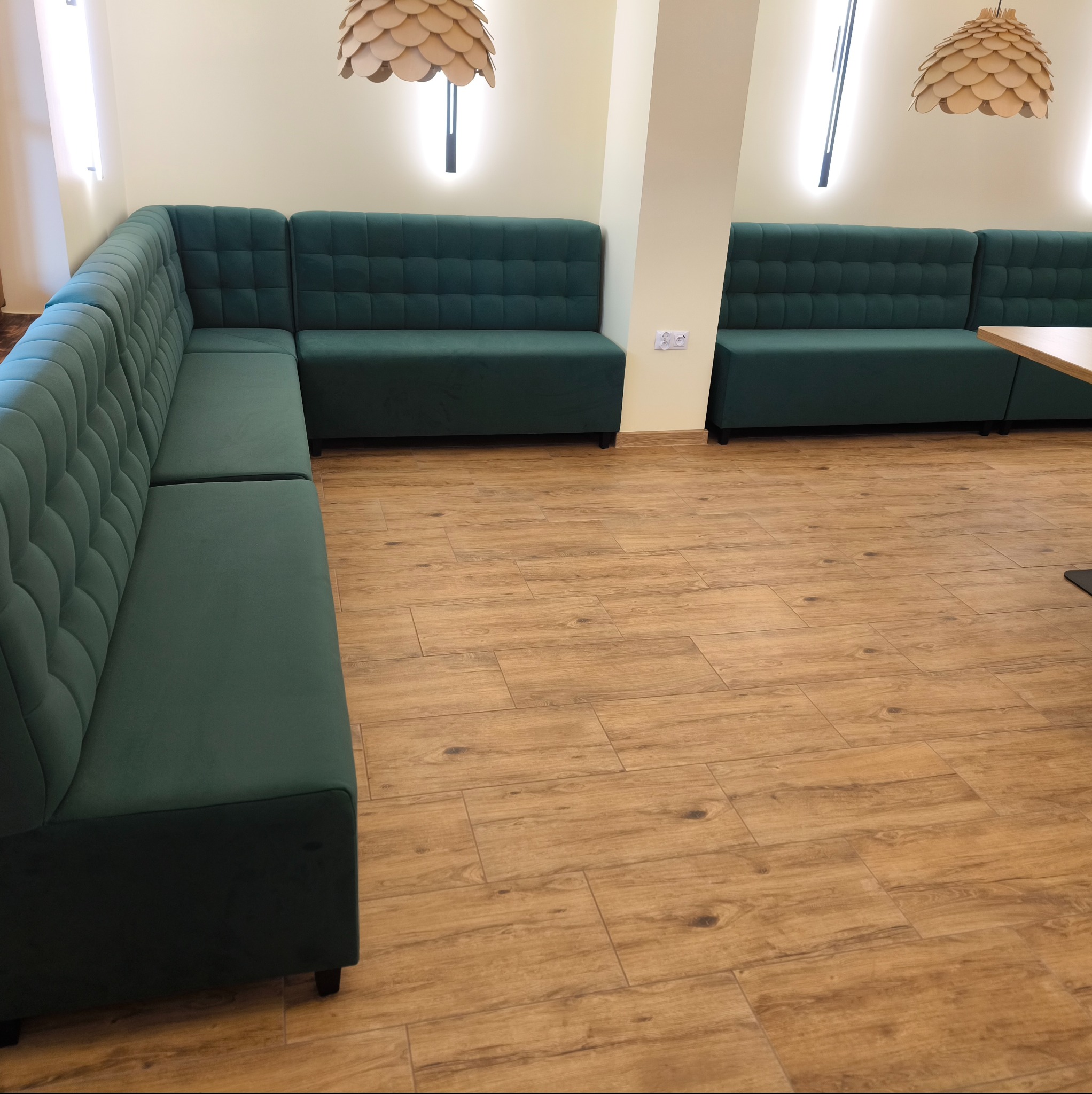 Długa, zielona, tapicerowana sofa z pikowanym wzorem w lokalu gastronomicznym. Drewnopodobna podłoga i ozdobne lampy nadają wnętrzu przytulny klimat.