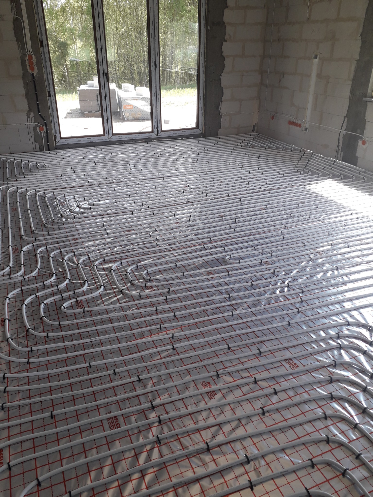 Rozłożona instalacja ogrzewania podłogowego z rur na folii z siatką w surowym stanie, widoczne okno z widokiem na zewnątrz, ściany z betonu komórkowego.