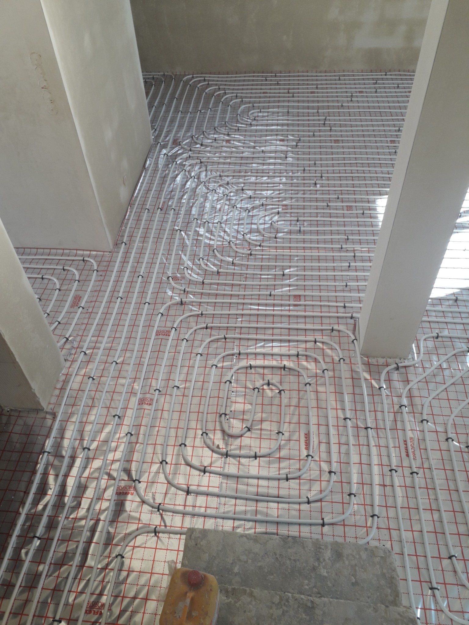 Rozłożona instalacja ogrzewania podłogowego na folii refleksyjnej z siatką, widoczne białe rury mocowane czarnymi uchwytami, surowy stan wykończenia z betonowymi filarami.