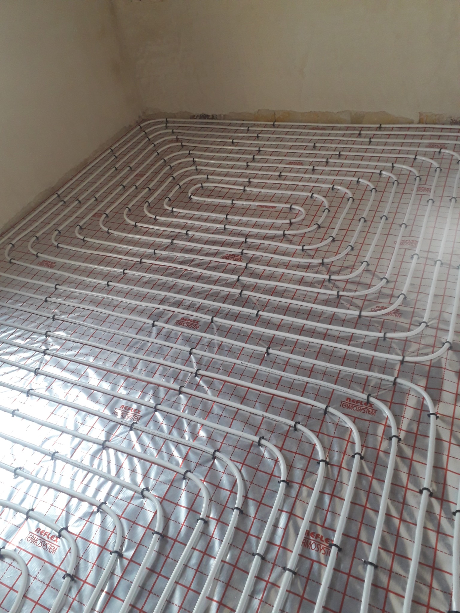 Białe rury ogrzewania podłogowego ułożone w spiralny wzór na folii z czerwoną siatką, widok z góry w rogu nieotynkowanego pomieszczenia.