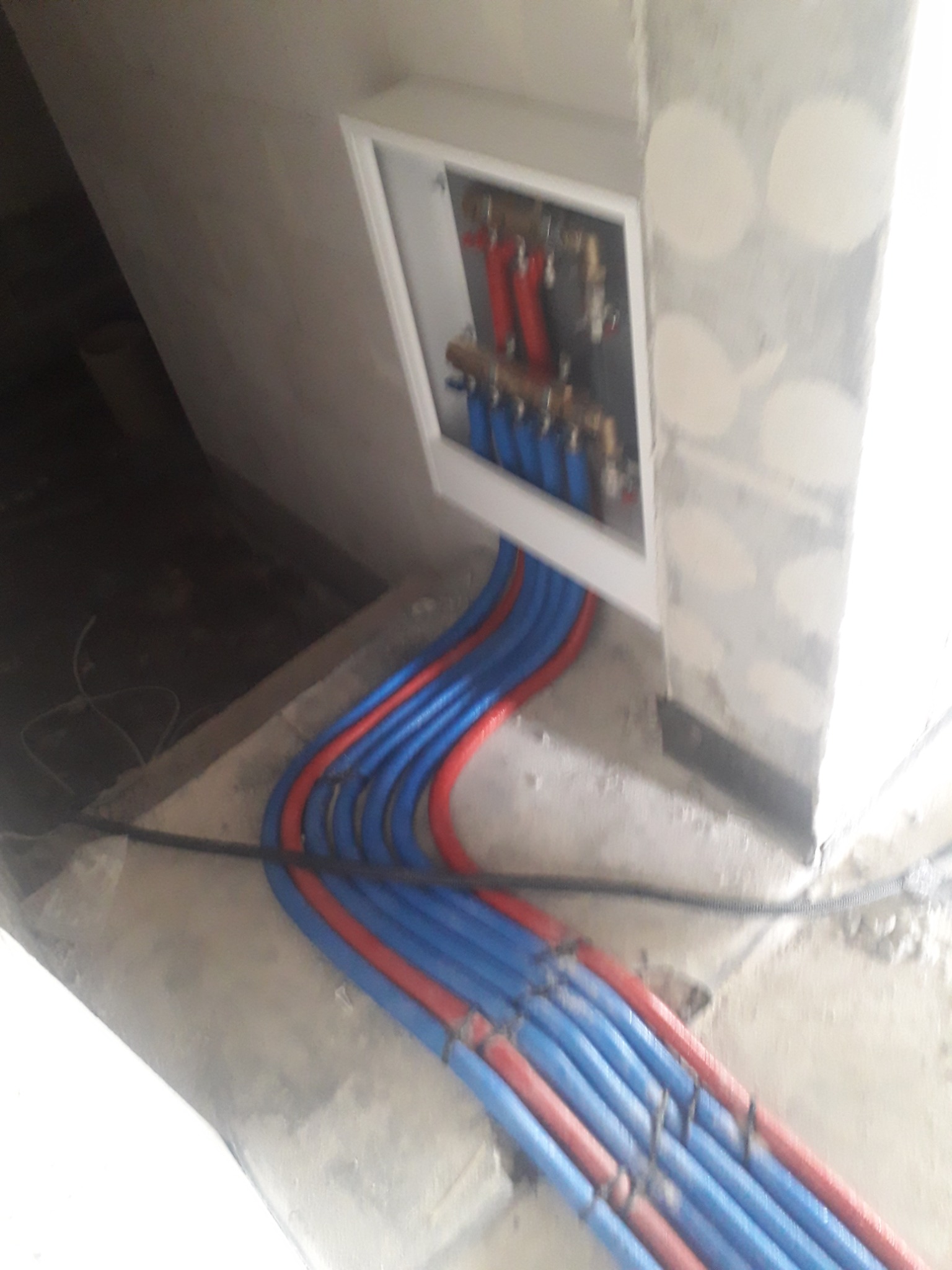 Rozdzielacz centralnego ogrzewania i ciepłej wody użytkowej z rurami PEX w kolorze niebieskim i czerwonym, umieszczony w białej szafce w surowym wnętrzu.