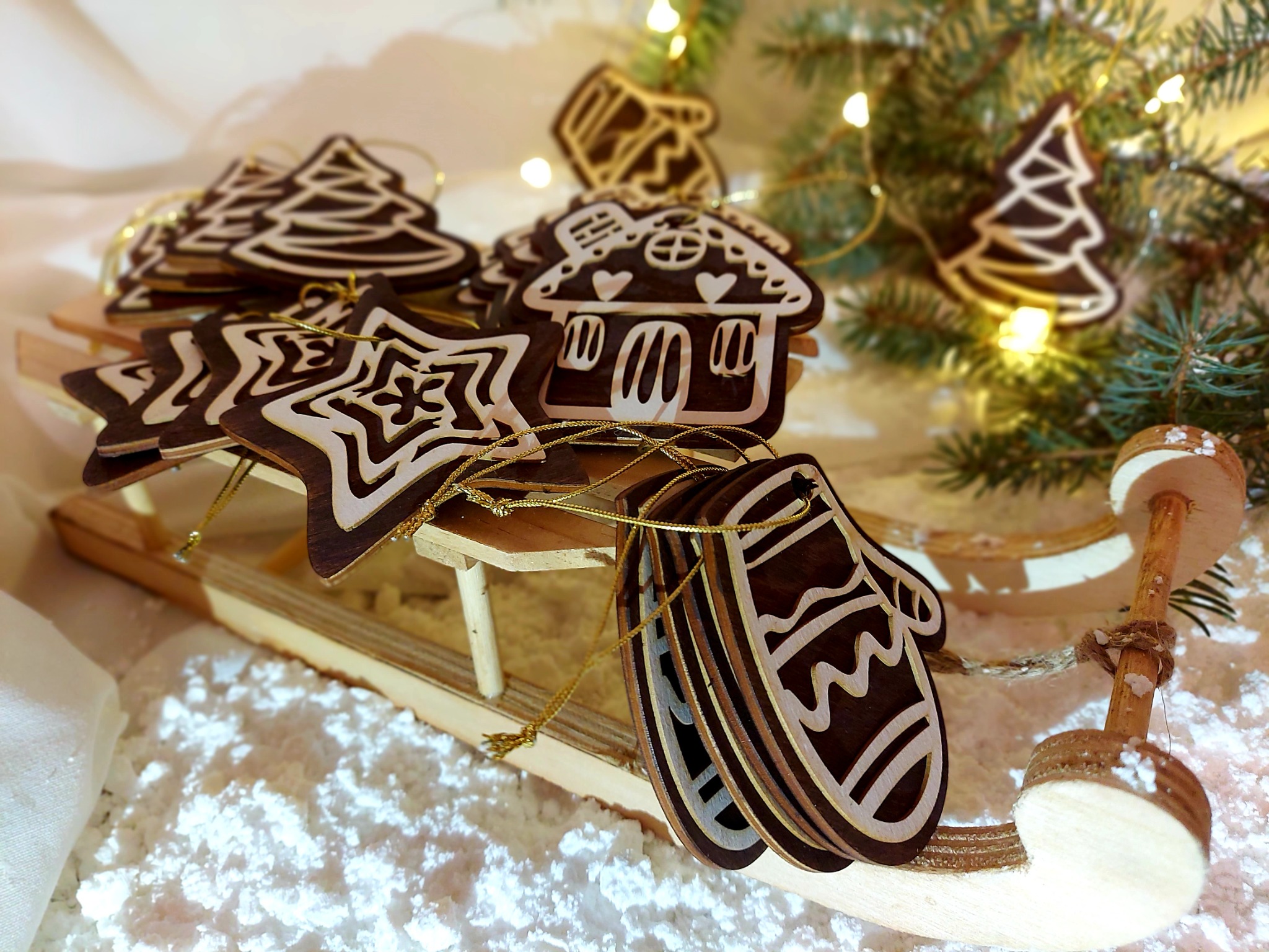 Ręcznie robione, drewniane ozdoby świąteczne w kształcie choinek, gwiazd, domków i rękawic, leżące na małych, drewnianych sankach, otoczone sztucznym śniegiem i gałązkami choinki, w ciepłym świetle.