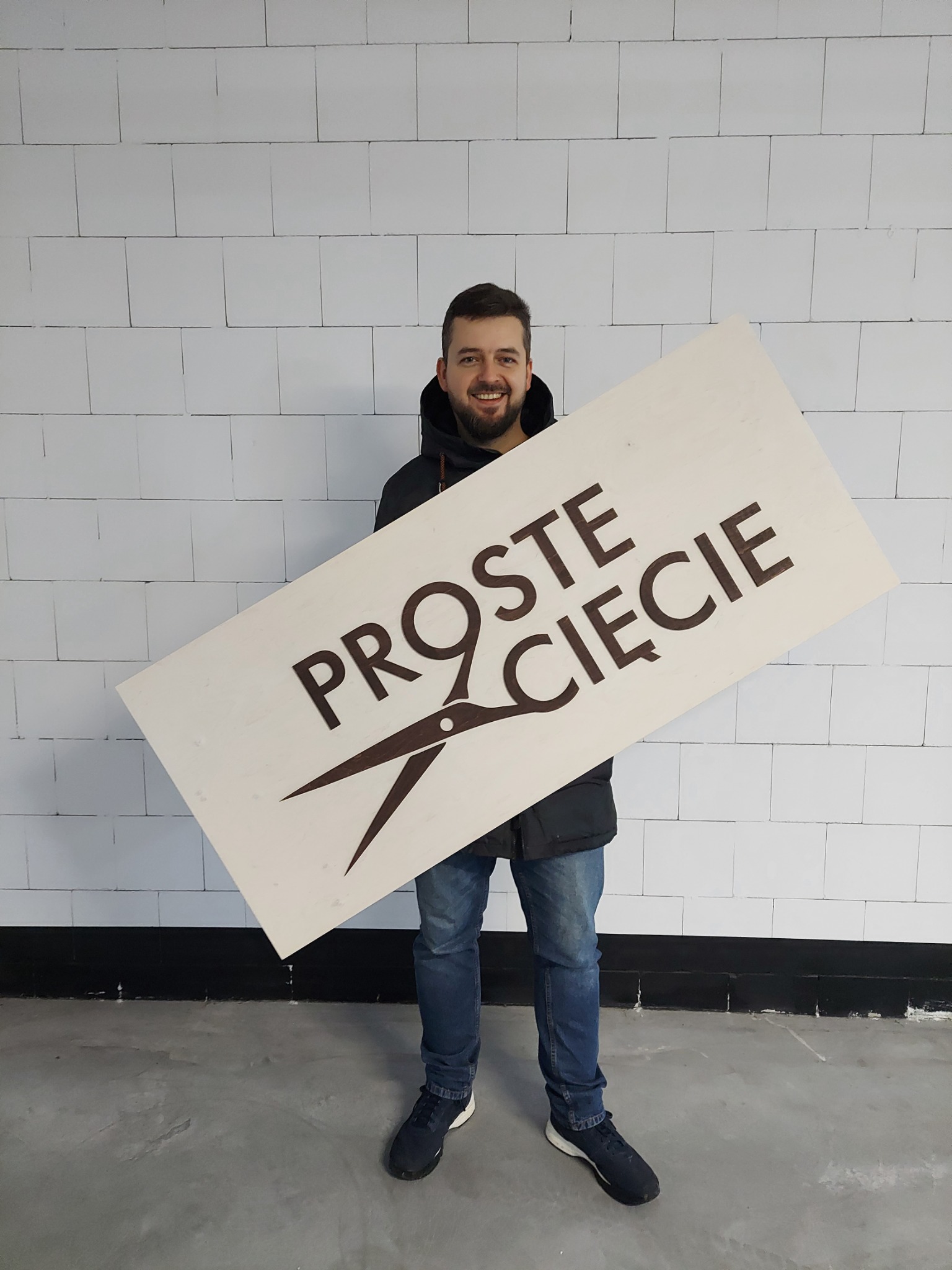 Uśmiechnięty mężczyzna trzymający dużą, prostokątną tablicę z napisem 'PROSTE CIĘCIE' i grafiką nożyczek, na tle ściany z białych cegieł.
