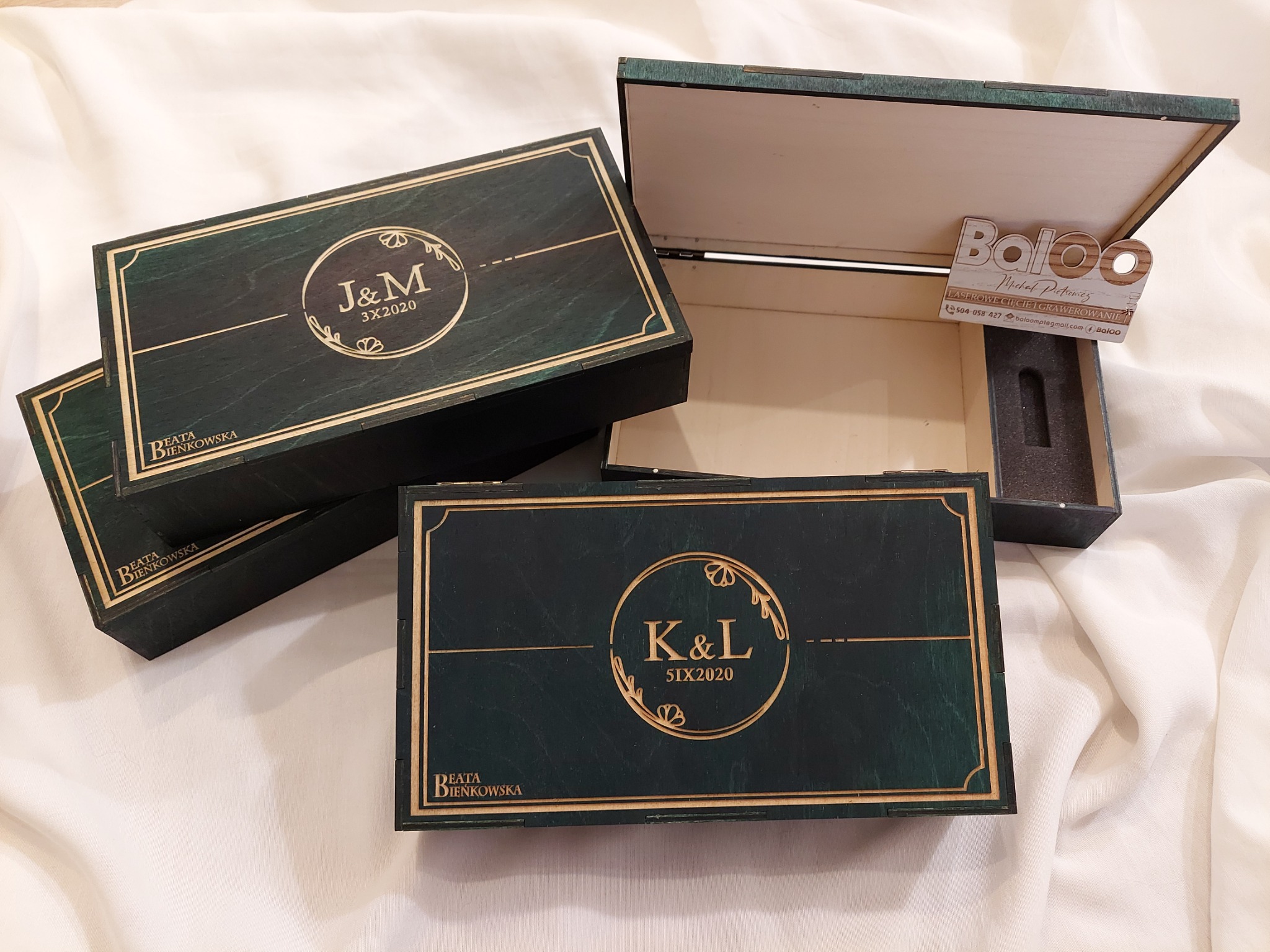 Drewniane, lakierowane pudełka z wygrawerowanymi inicjałami i datą, ułożone na białym materiale. Na jednym z pudełek widać logo firmy Baloo.