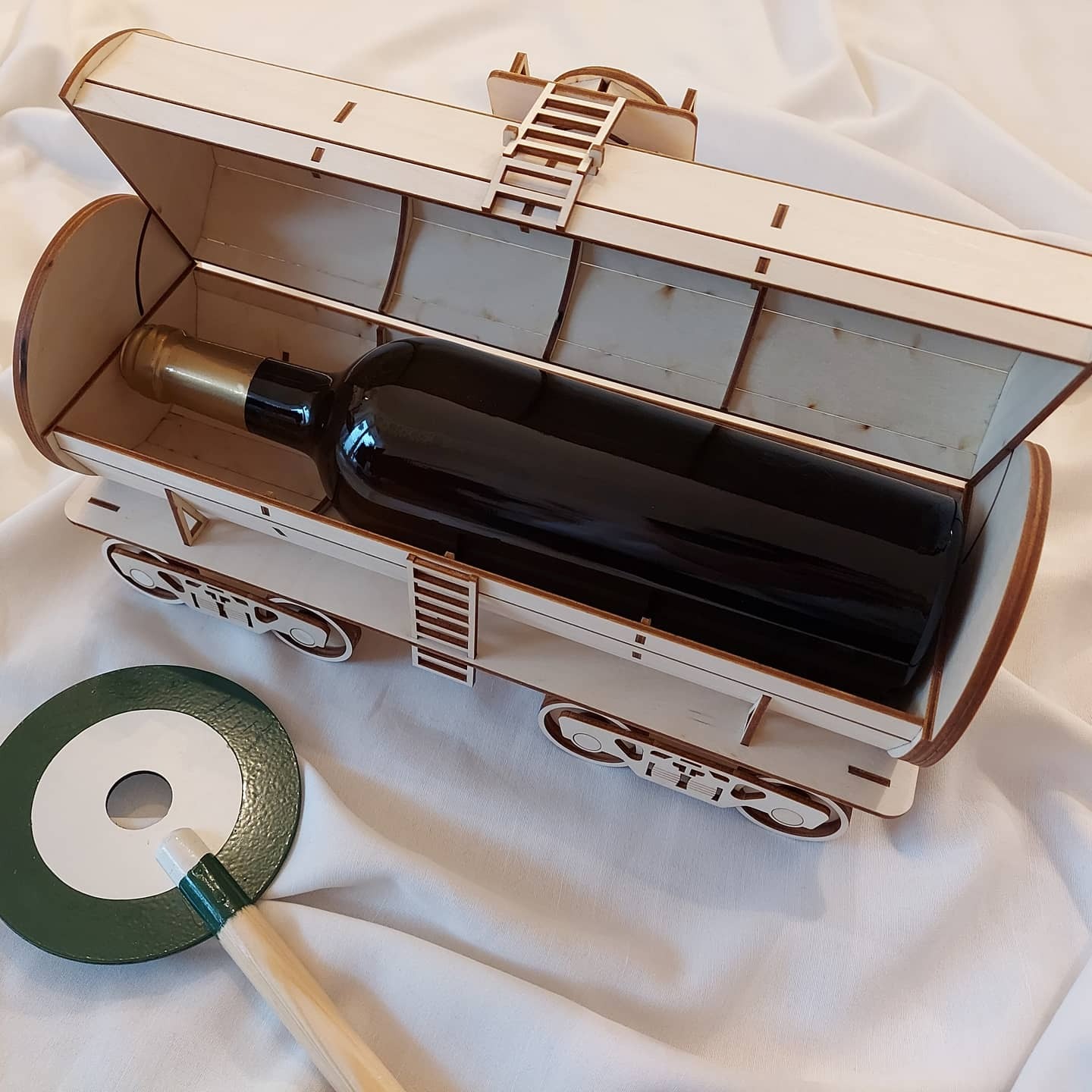 Drewniany wagonik z otwieraną klapą, zawierający butelkę wina, obok leży okrągły, zielono-biały podkładka pod kieliszek z drewnianą rączką, całość na białym tle.