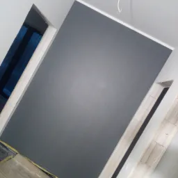 Świeżo pomalowana ściana na szaro, widoczne fragmenty futryn drzwiowych i zabezpieczona podłoga, kabel zwisający z sufitu.
