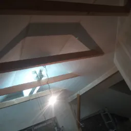 Skośny sufit poddasza z drewnianą konstrukcją szkieletową, przygotowany do ocieplenia, z widocznym oknem dachowym i punktowym oświetleniem.