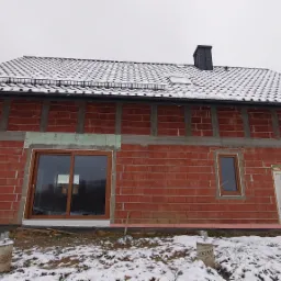 Surowy budynek z czerwonej cegły w trakcie budowy, z zamontowanymi oknami z brązowymi ramami i białą, prowizoryczną stolarką drzwiową, pokryty śniegiem.