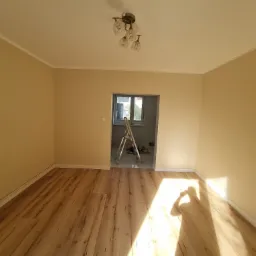 Pokój po remoncie, świeżo pomalowane ściany w odcieniach beżu, panele podłogowe imitujące drewno, widoczna drabina w przejściu do innego pomieszczenia, lampa sufitowa z kilkoma szklanymi kloszami.