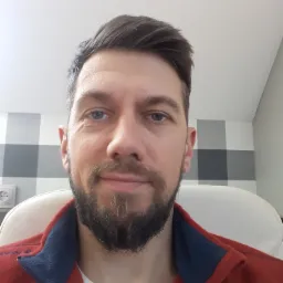 Portret brodatego mężczyzny w czerwonej bluzie na tle ściany z szaro-białą kratą i białego krzesła.