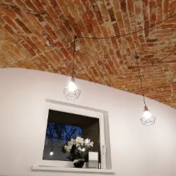 Wnętrze z ceglanym sklepieniem łukowym, trzema wiszącymi lampami z geometrycznymi kloszami na czarnych kablach, białymi ścianami i oknem z widokiem na drzewa oświetlone wieczornym światłem,...