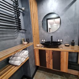 AllBud Usługi Ogólnobudowlane Łokietek Kamil - Nowoczesna łazienka z szarą ścianą o fakturze betonu, czarną umywalką w kształcie diamentu, okrągłym lustrem, drewnianymi elementami wykończeniowymi i czarnym grzejnikiem drabinkowym. Na półce leżą...