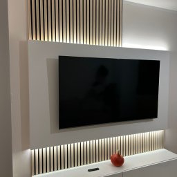 AllBud Usługi Ogólnobudowlane Łokietek Kamil - Ściana telewizyjna z podświetlanymi lamelami drewnianymi, biała szafka RTV z pomarańczową dekoracją w kształcie dyni.