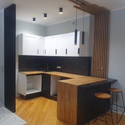 AllBud Usługi Ogólnobudowlane Łokietek Kamil - Nowoczesna kuchnia z drewnianym blatem, czarnymi elementami i białymi szafkami, uzupełniona drewnianymi lamelami na ścianie i minimalistycznymi lampami wiszącymi.