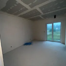 Pokój w stanie deweloperskim: surowe ściany, podłoga z wylewki, sufit z płyt kartonowo-gipsowych ze spoinami, okno balkonowe z widokiem na zewnątrz, niebieska wiązka kabli na podłodze.