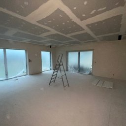 AllBud Usługi Ogólnobudowlane Łokietek Kamil - Pokój w trakcie prac wykończeniowych: ściany i sufit po szpachlowaniu, widoczne łączenia płyt karton-gips na suficie, drabina aluminiowa na środku, okna zabezpieczone folią malarską.