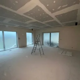 Pokój w trakcie prac wykończeniowych: ściany i sufit po szpachlowaniu, widoczne łączenia płyt karton-gips na suficie, drabina aluminiowa na środku, okna zabezpieczone folią malarską.